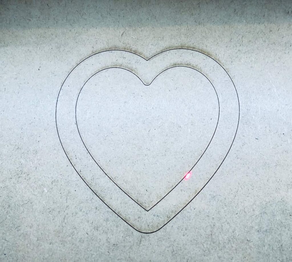 Image shows mdf board with laser cut heart being cut out.