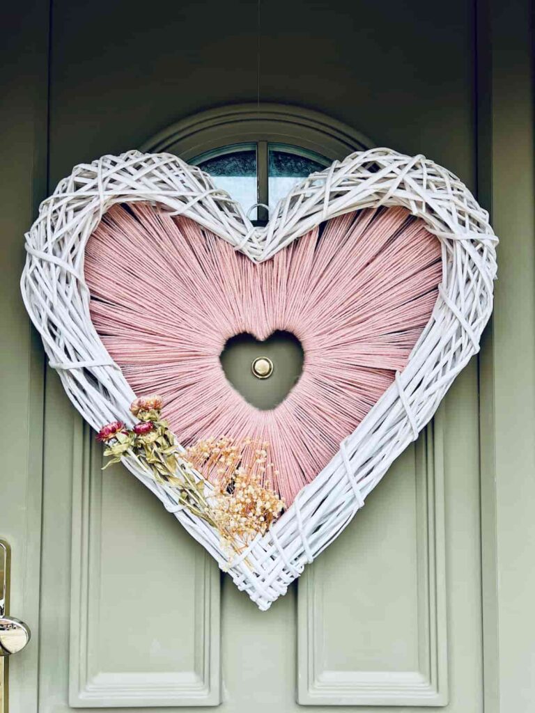 Image shows yarn heart shaped wreath with faux florals on a green front door.