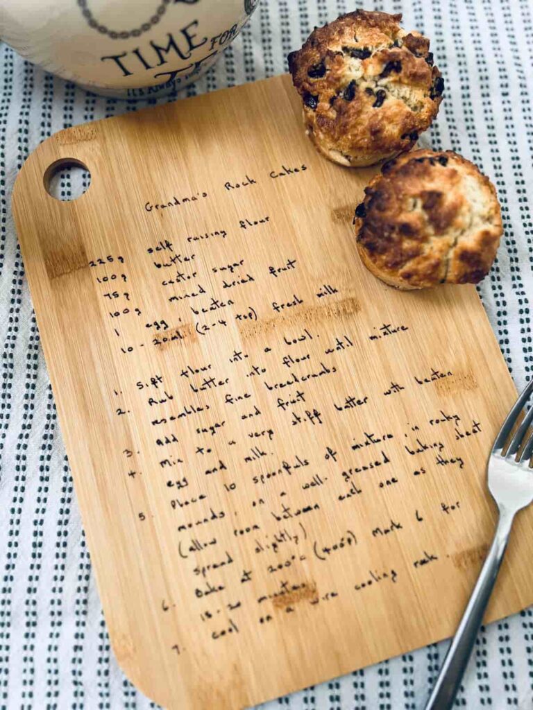 image shows chopping board with engraved recipe. Plus cakes, a fork and teapot around the edges.