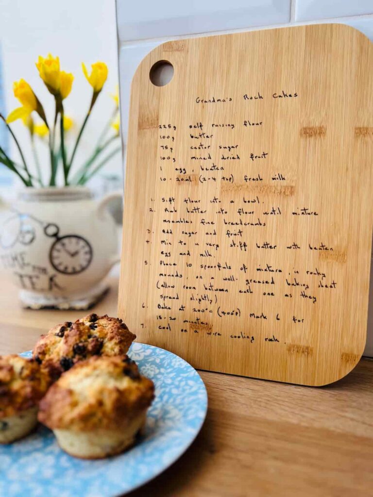 image shows chopping board with egrnaved recipe and cakes on a plate and flowers in the background.