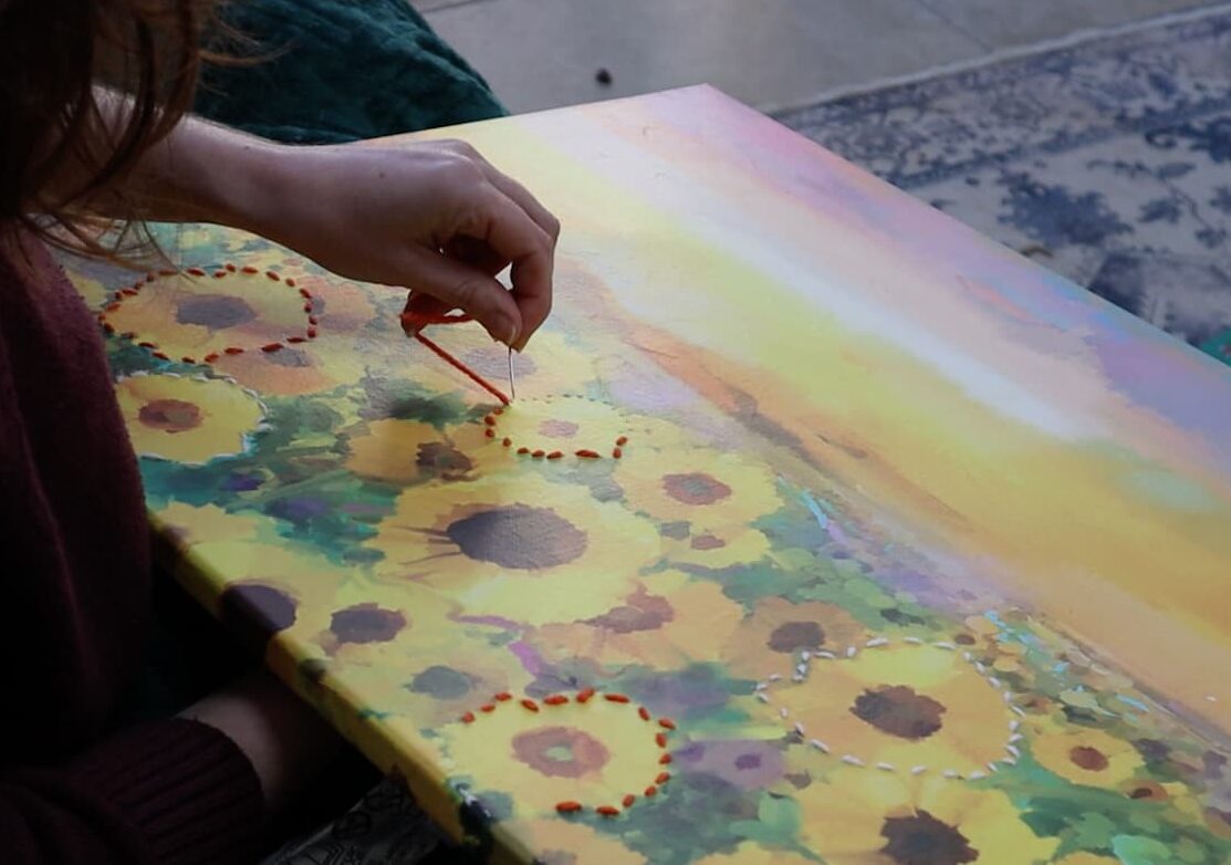 image shows Rachel sitting down while doing embroidery on the canvas.